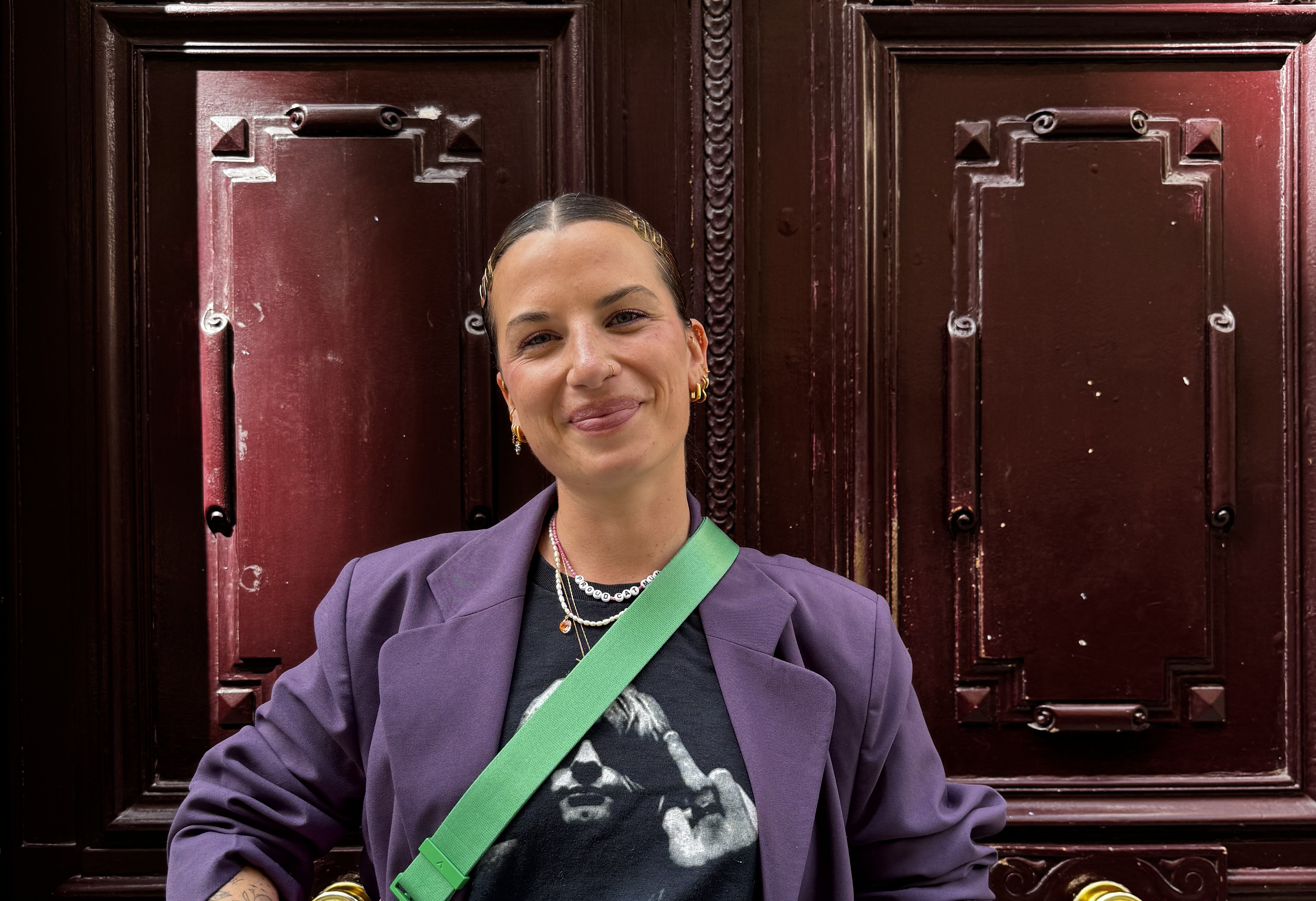 Elena devant une porte dans la rue, elle porte un sac en bandoulière et une mini jupe.