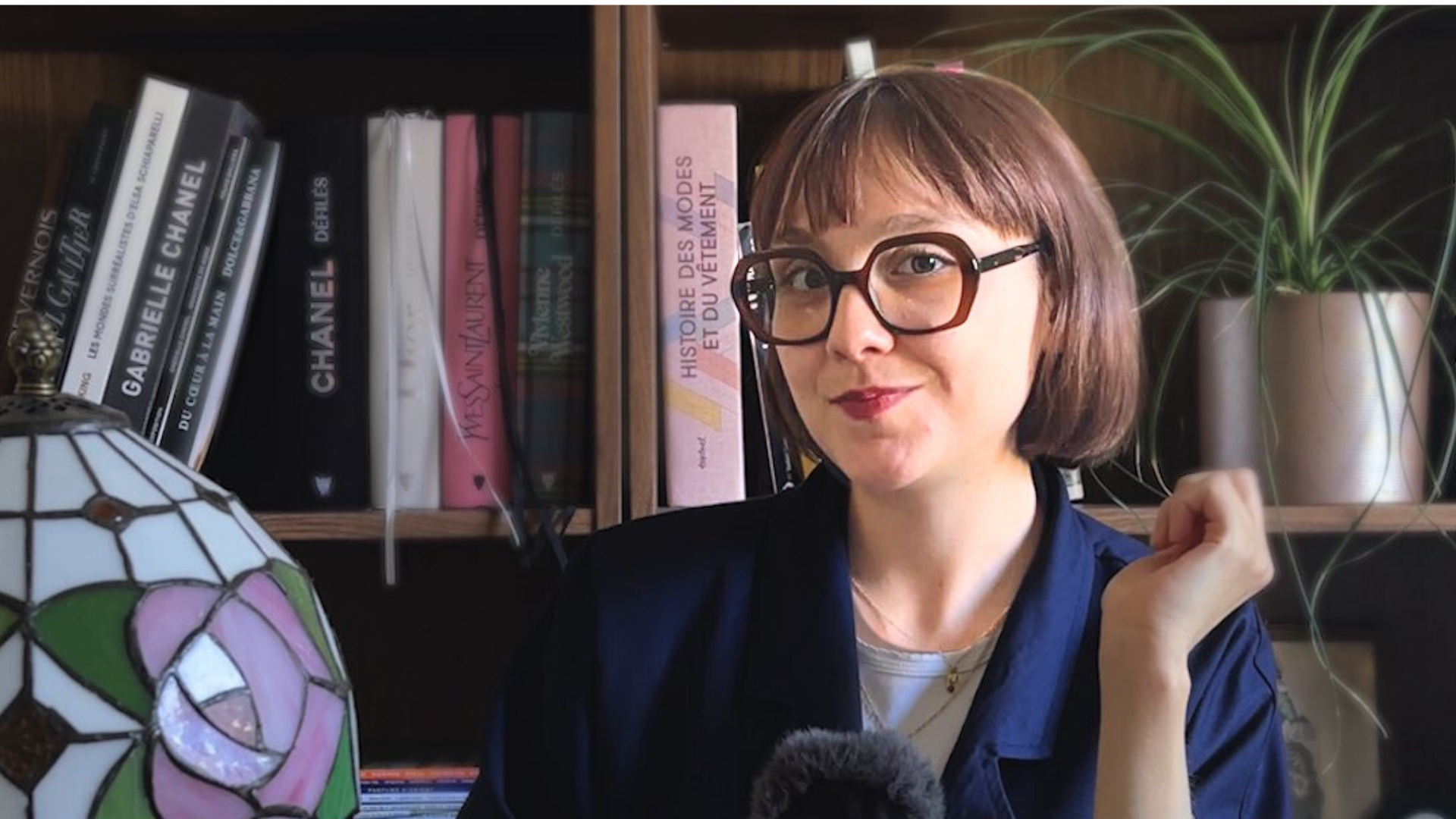 Clara a un micro à la main et est assise devant sa blibliothèque, une jolie lampe arts-déco sur le bureau